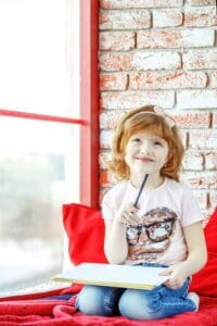 A beautiful kid thinks and draws. The girl is sitting on the win